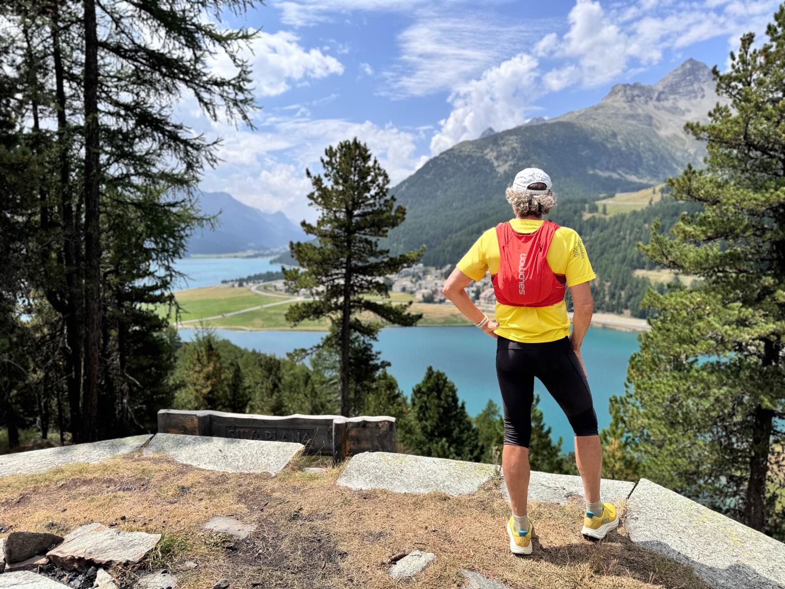 Vista panoramica del Lago di Silvaplana in Alta Engadina con escursionista seduto su una panchina tra i pini e le montagne svizzere.